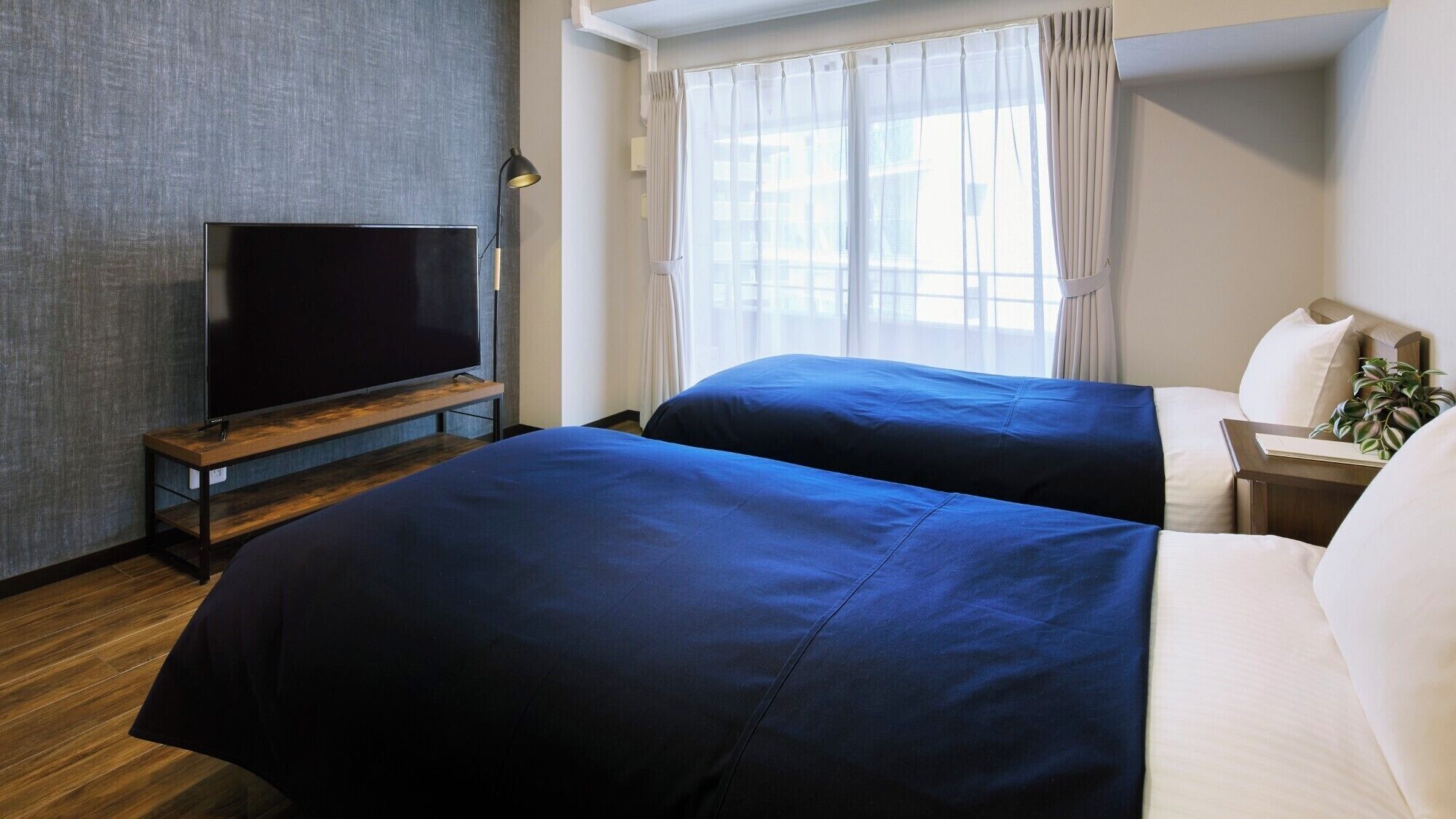 Een hotelkamer met twee bedden, een televisie op een houten standaard, een nachtkastje met een lamp en een raam met dunne gordijnen.
