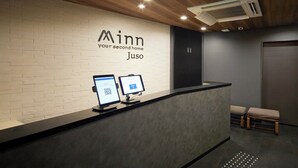 A reception desk with a white brick wall and a logo that reads "Minn your second home Juso". There are two computer monitors on the desk, and a wooden bench is visible in the background. - Minn Juso (Osaka)