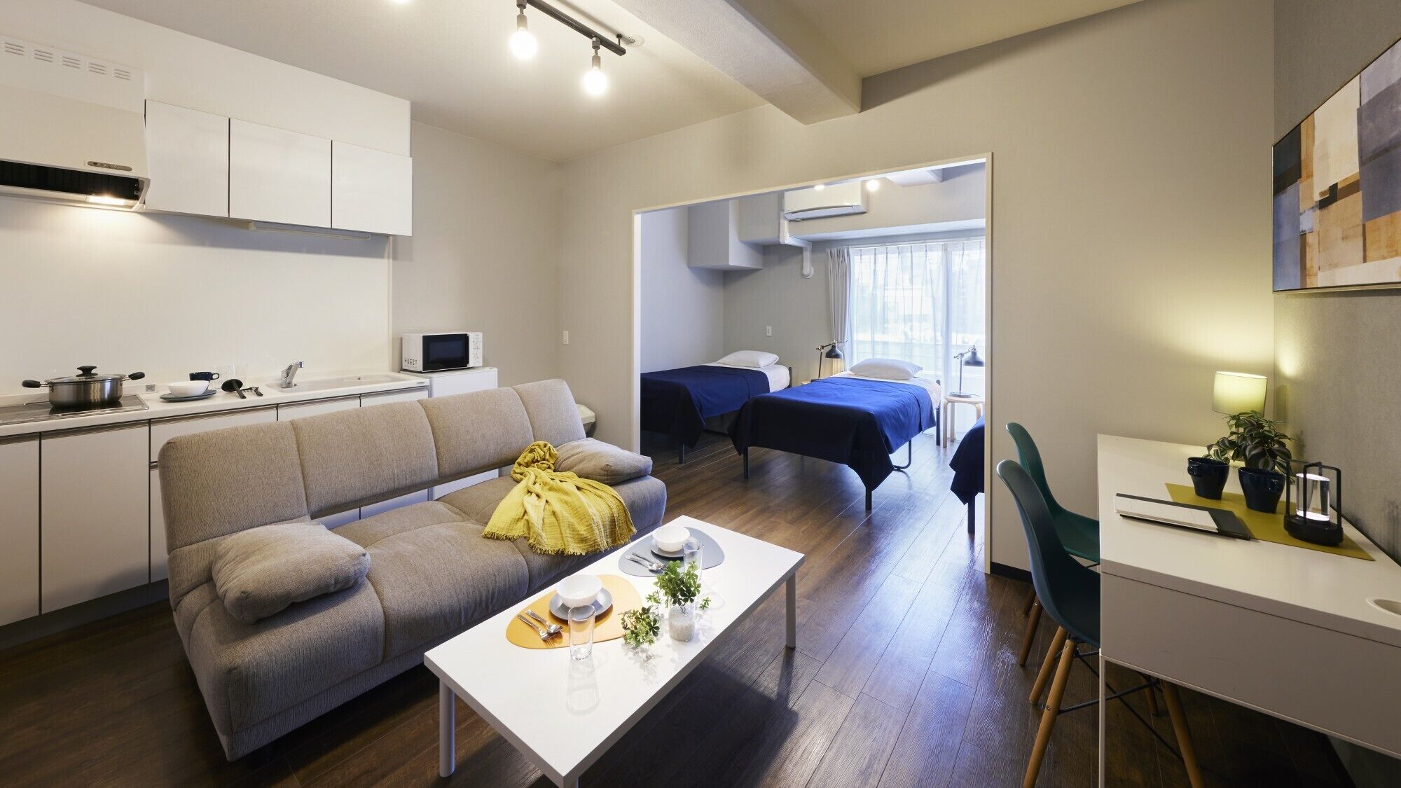 A modern apartment with a kitchen, a living area with a sofa, a dining table, two beds, and a small desk.