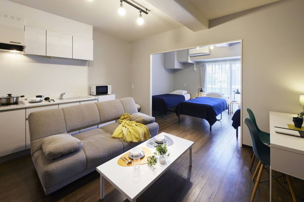 A modern apartment with a kitchen, a living area with a sofa, a dining table, two beds, and a small desk.
