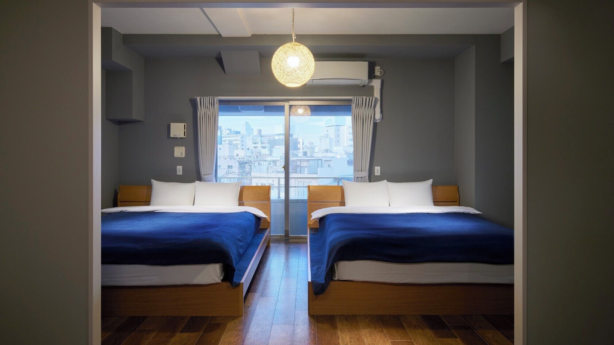 A room with two beds, a large window with curtains, a hanging light fixture, and a view of the cityscape.