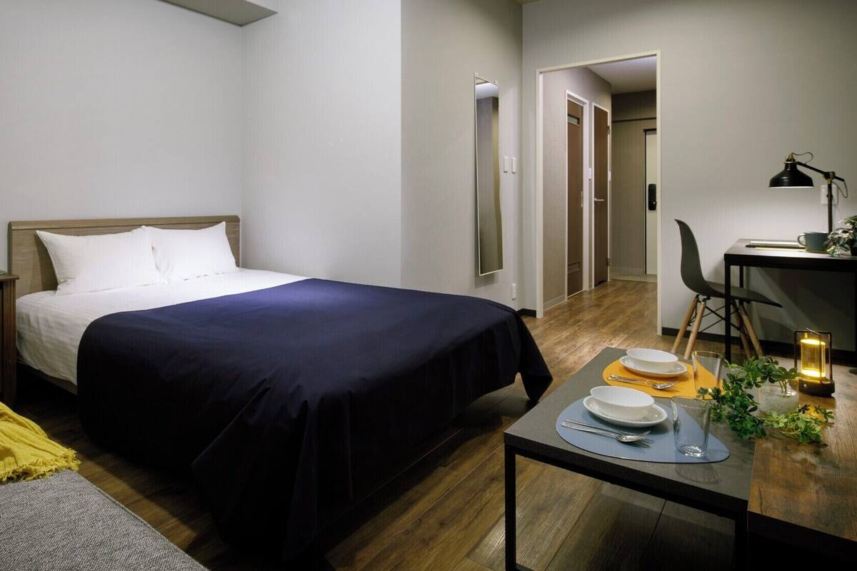 A hotel room with a bed, bedside table, chair, and a small desk with a lamp and a plant.