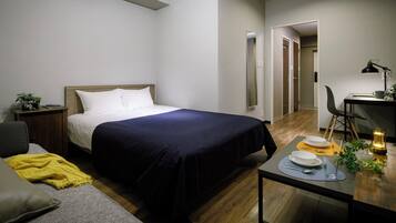 A hotel room with a bed, bedside table, chair, and a small desk with a lamp and a plant.