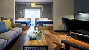A modern hotel room with a sofa, a small table with a bowl and glasses, a desk with a lamp, and two beds with blue bedding. - Minn Juso (Osaka)