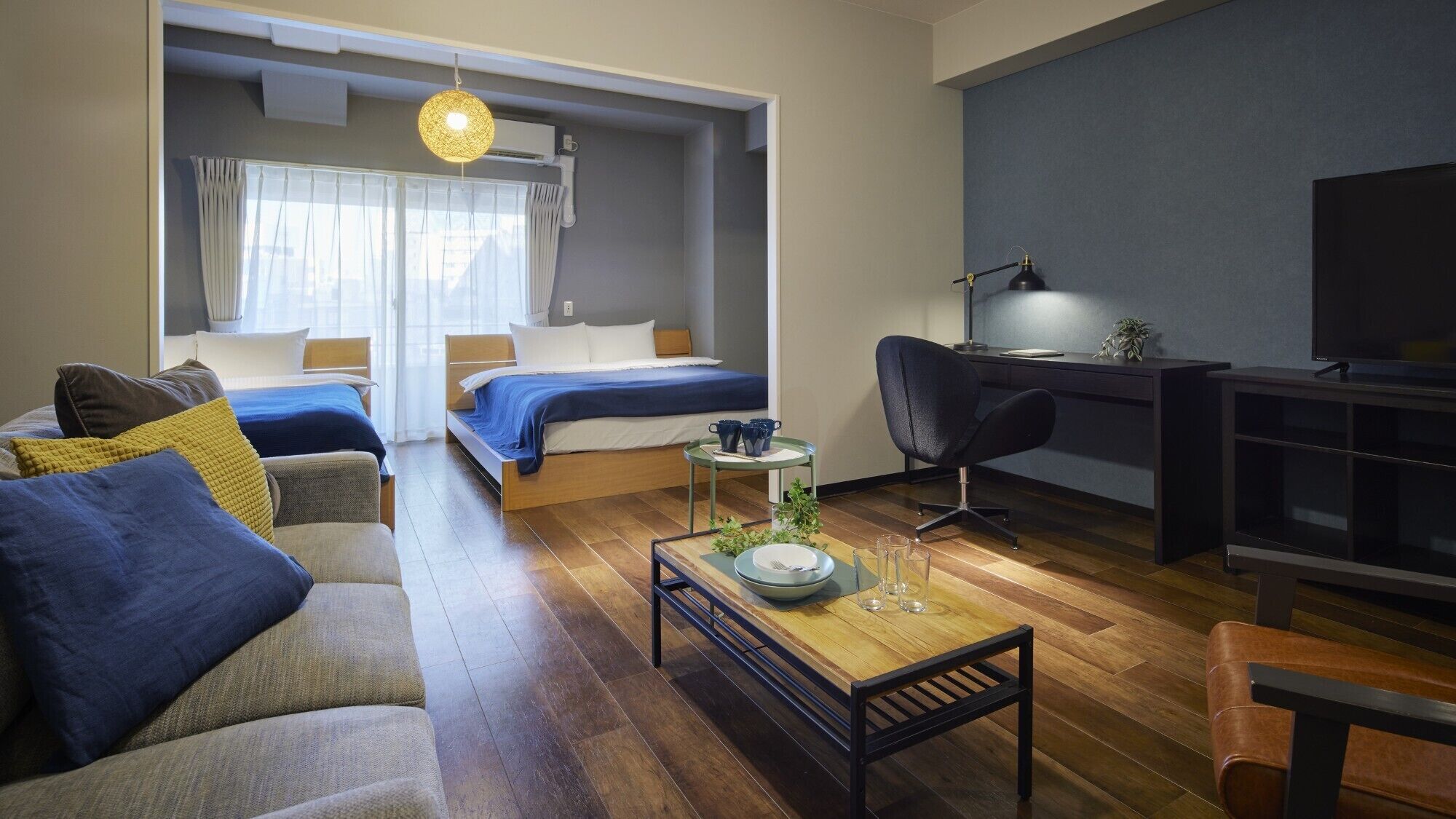 A modern hotel room with a sofa, two beds, a desk, a TV, and a dining table.