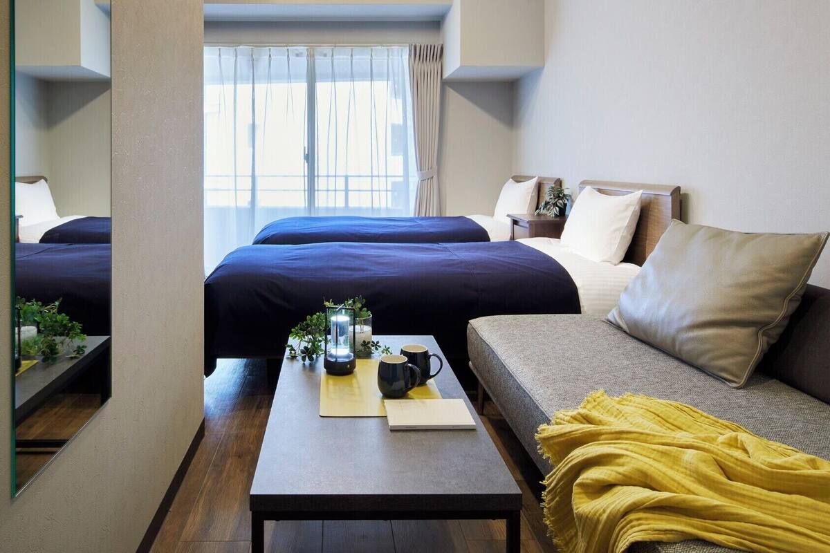 A hotel room with two beds, a sofa, a small table with a lamp, and a window with curtains.
