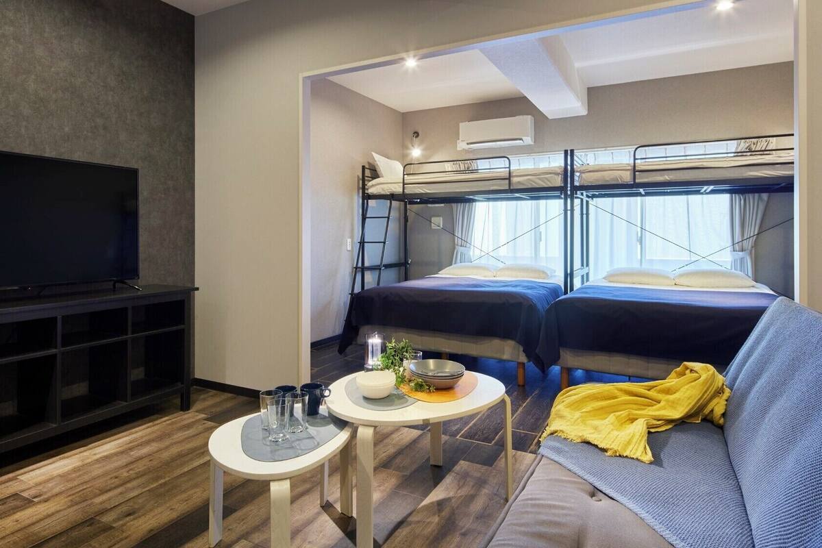 A compact hotel room with a TV, bunk beds, a sofa, and a small table with glasses and a bowl.