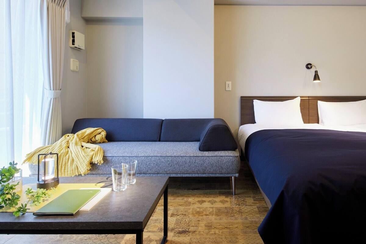 A hotel room with a sofa, a bed, a bedside table with glasses, a lamp, and a window with curtains.