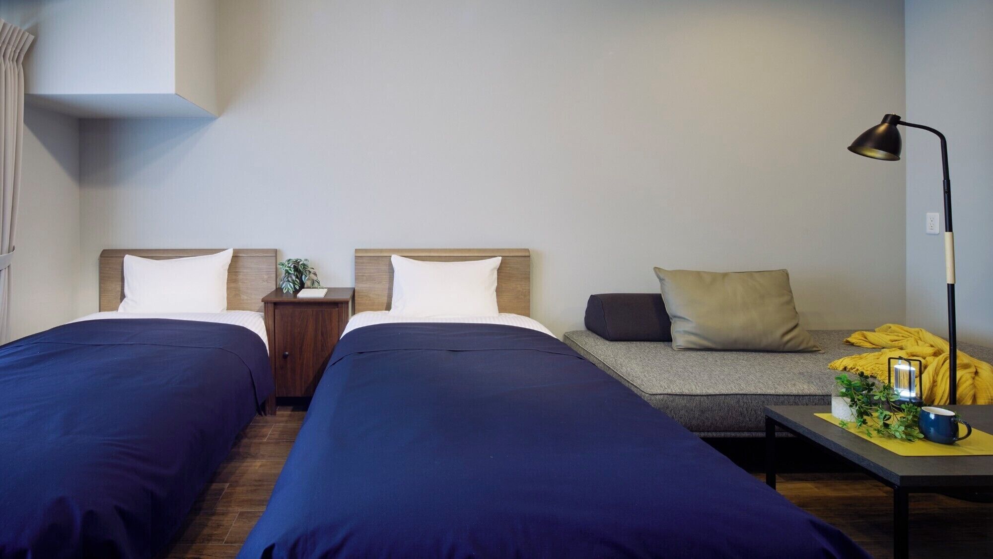 A hotel room with two beds, a nightstand, a sofa, a lamp, and a small table with a plant and a cup.