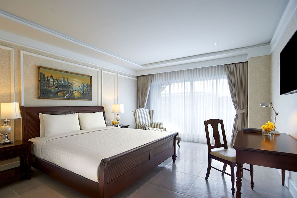 A hotel room with a large bed, a desk, a chair, a painting on the wall, and a TV on the wall.