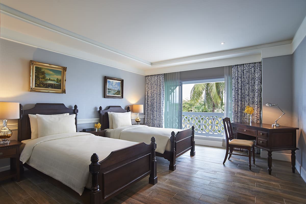 A hotel room with two beds, a desk, and a chair. There is a window with curtains and a view of palm trees.