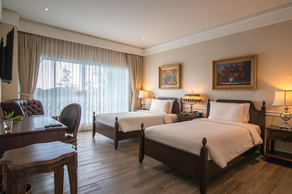 A hotel room with two beds, a desk, a chair, a TV, and artwork on the walls.