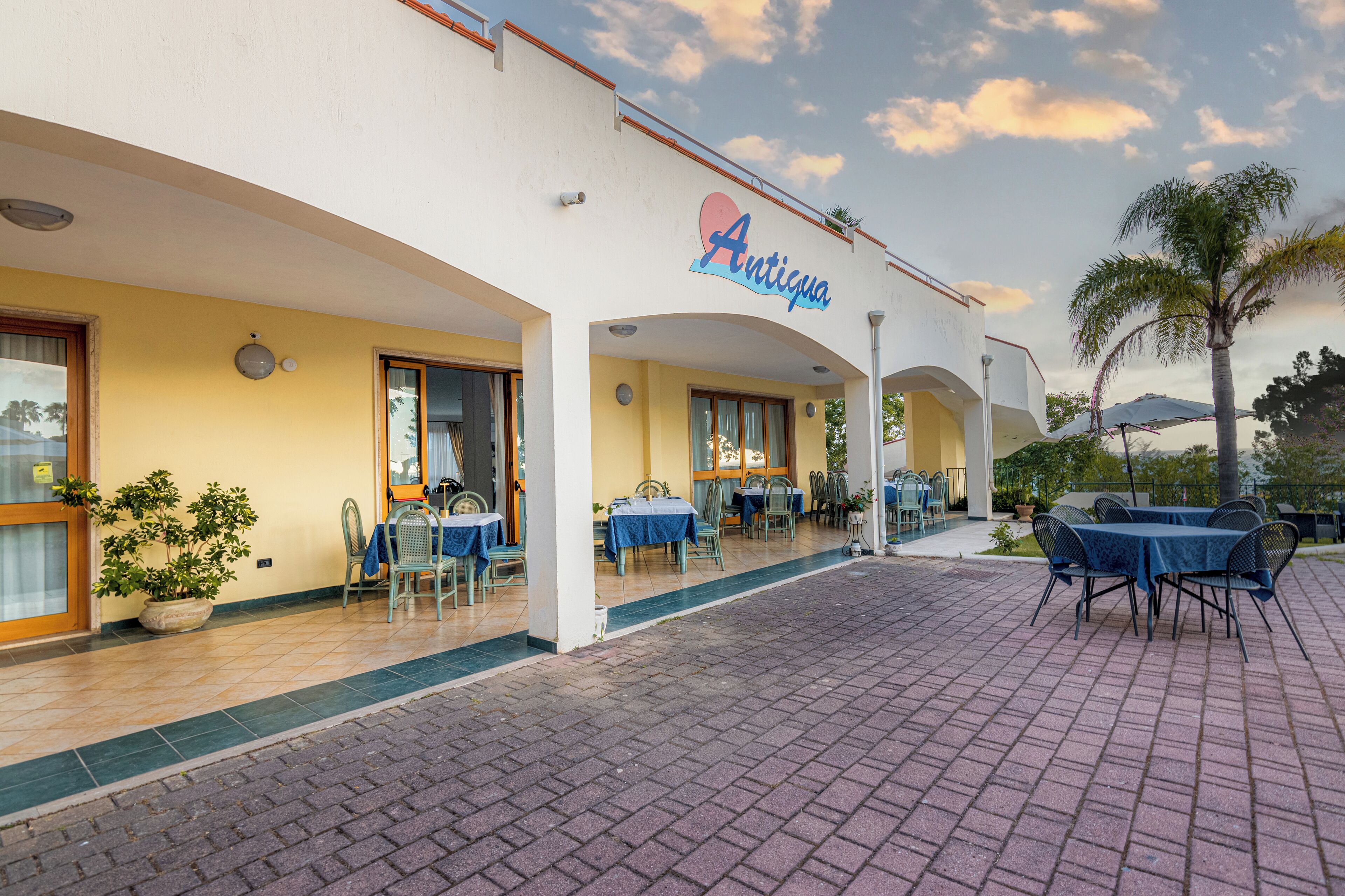 A restaurant named 'Antigua' with outdoor seating, potted plants, and a clear sky.