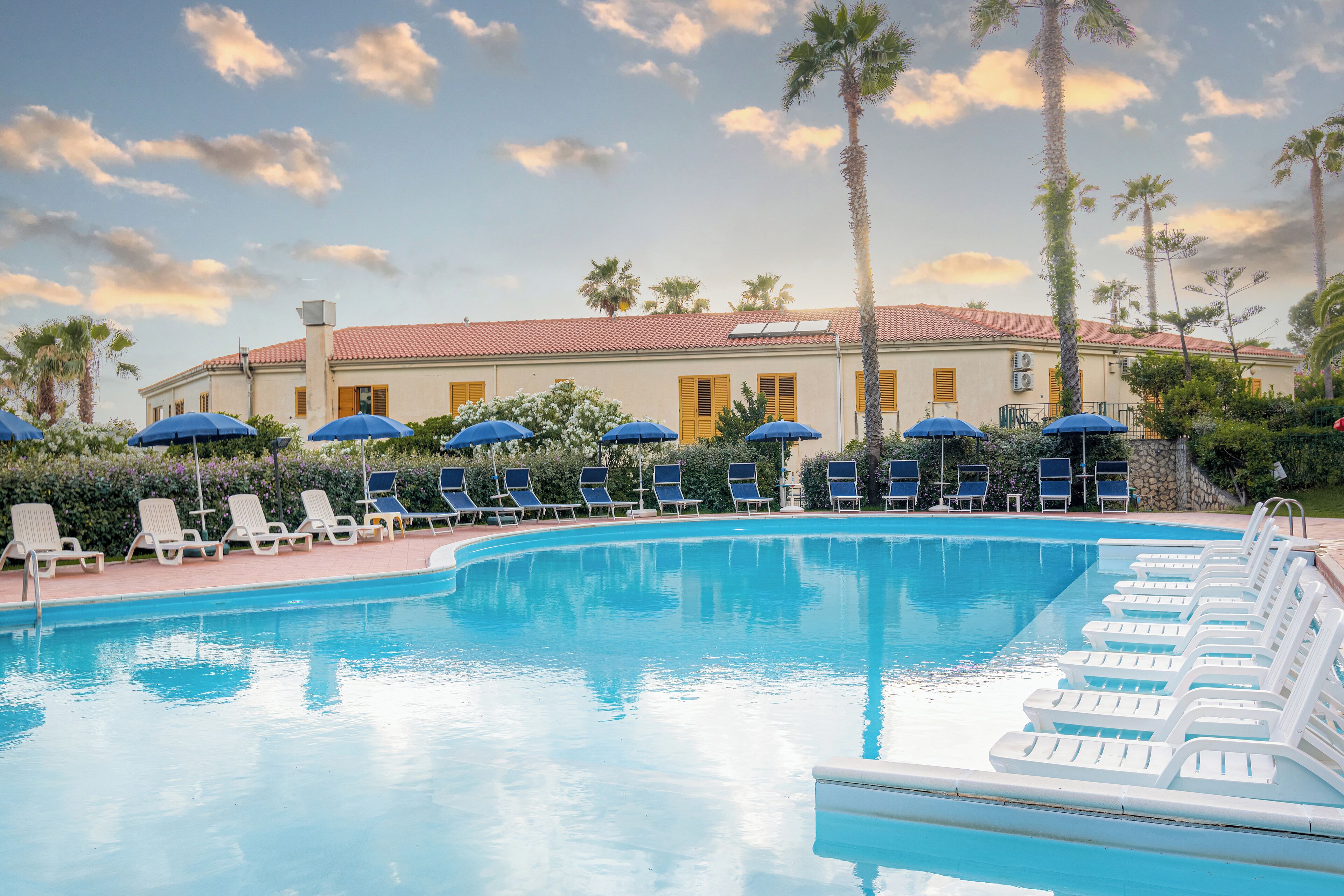 A swimming pool with lounge chairs and umbrellas, surrounded by palm trees and a building with a red-tiled roof.