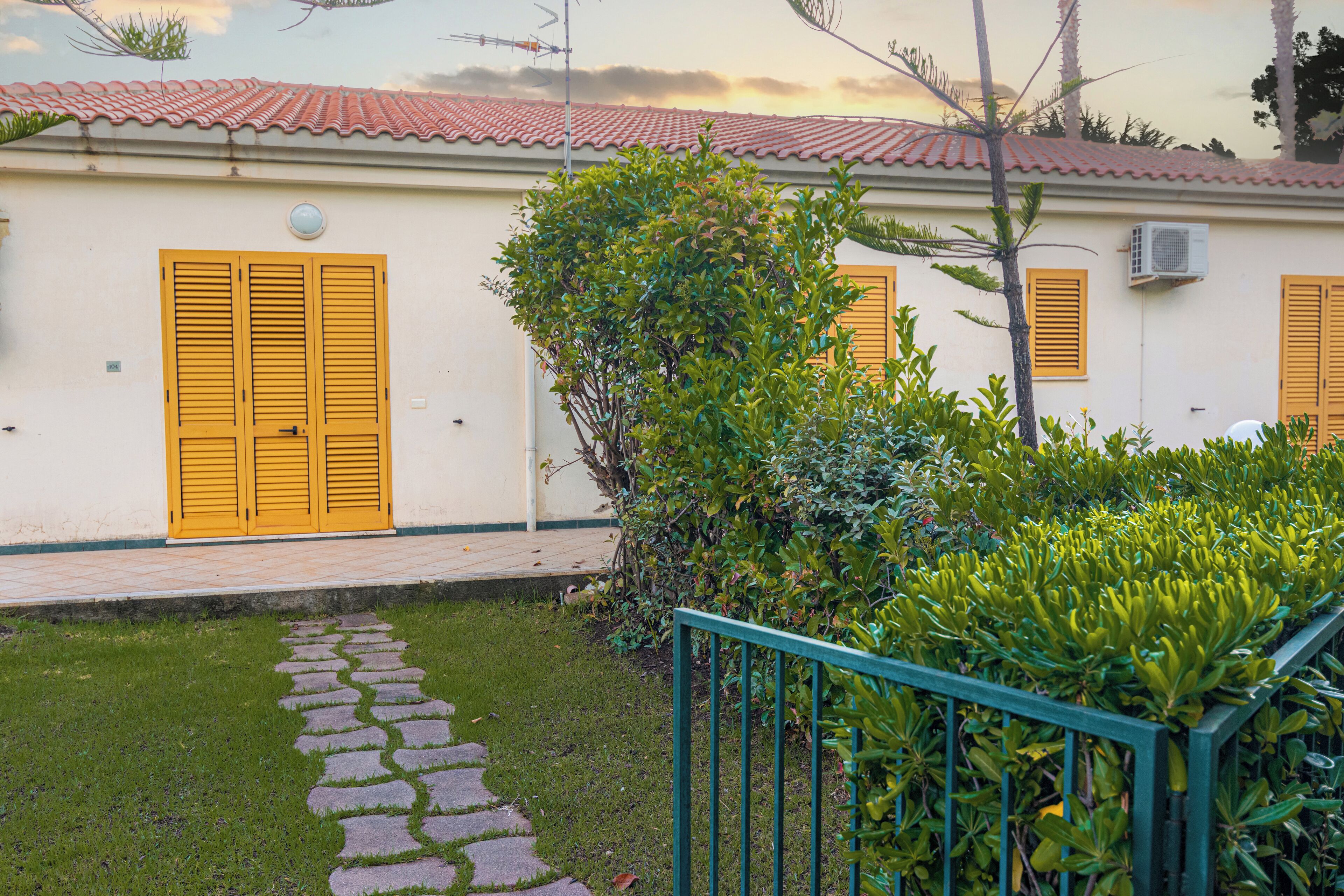 A building with yellow shutters, a tiled roof, and a paved pathway leading to the entrance.