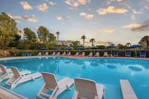 A swimming pool with white lounge chairs and blue umbrellas, surrounded by greenery and palm trees. - Residence Antigua (Bonifati)