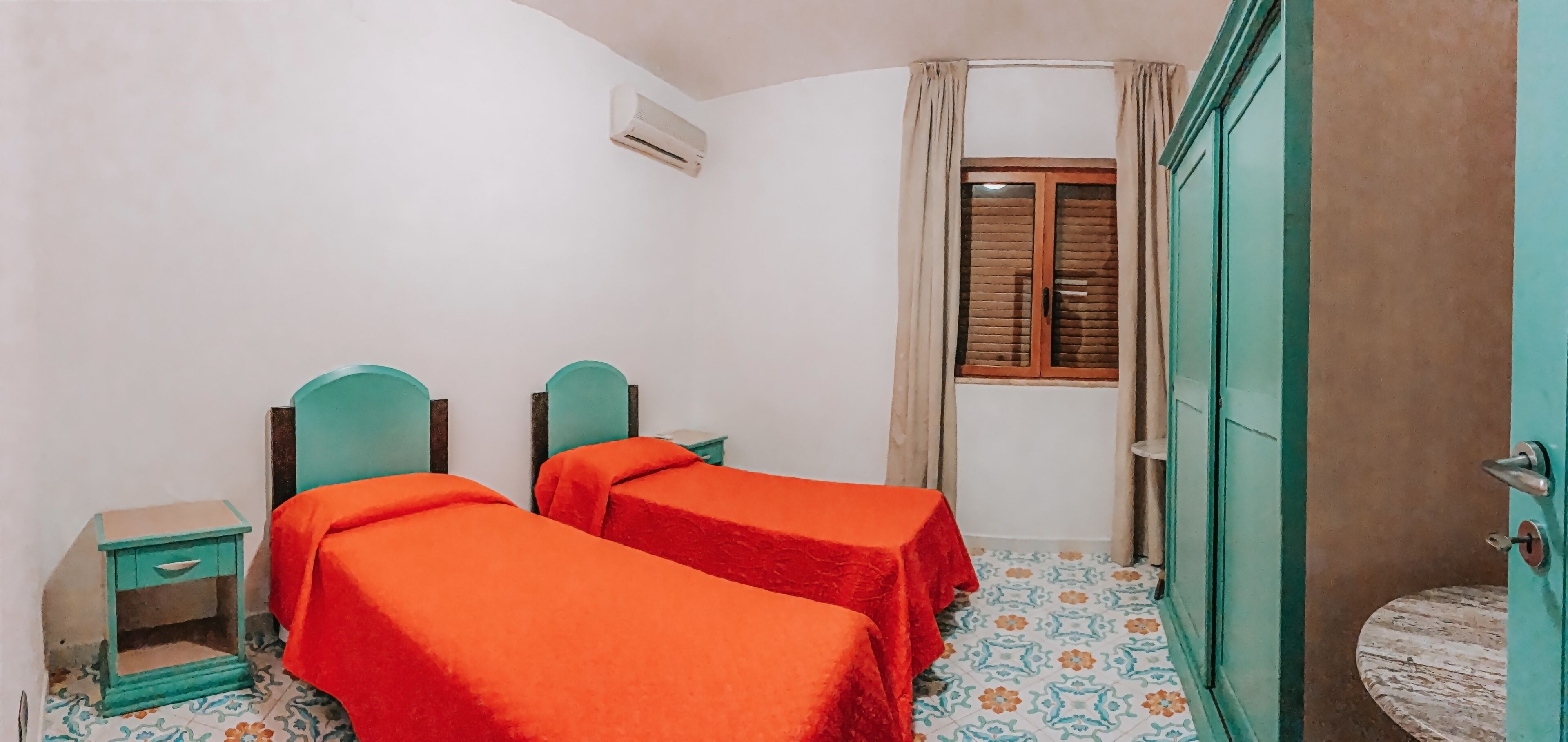 Quarto com duas camas, uma mesa pequena, uma mesa de cabeceira, uma janela com cortinas e uma porta.