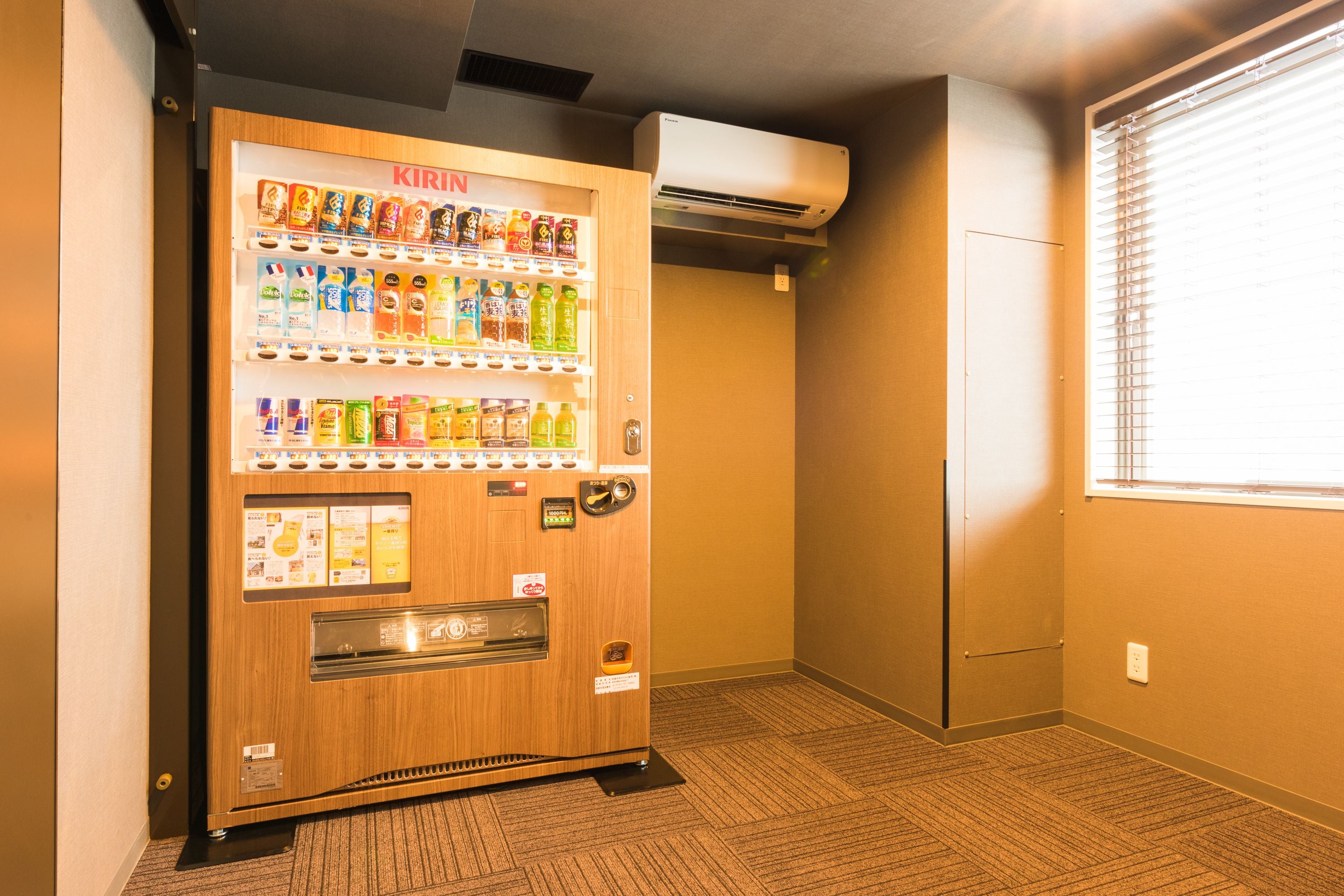 A vending machine with various beverages and snacks, located in a hallway with carpeted flooring and a window with blinds.
