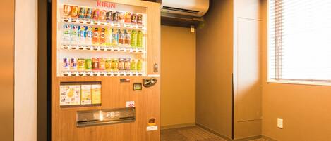 A vending machine with various beverages and snacks, located in a hallway with carpeted flooring and a window with blinds.