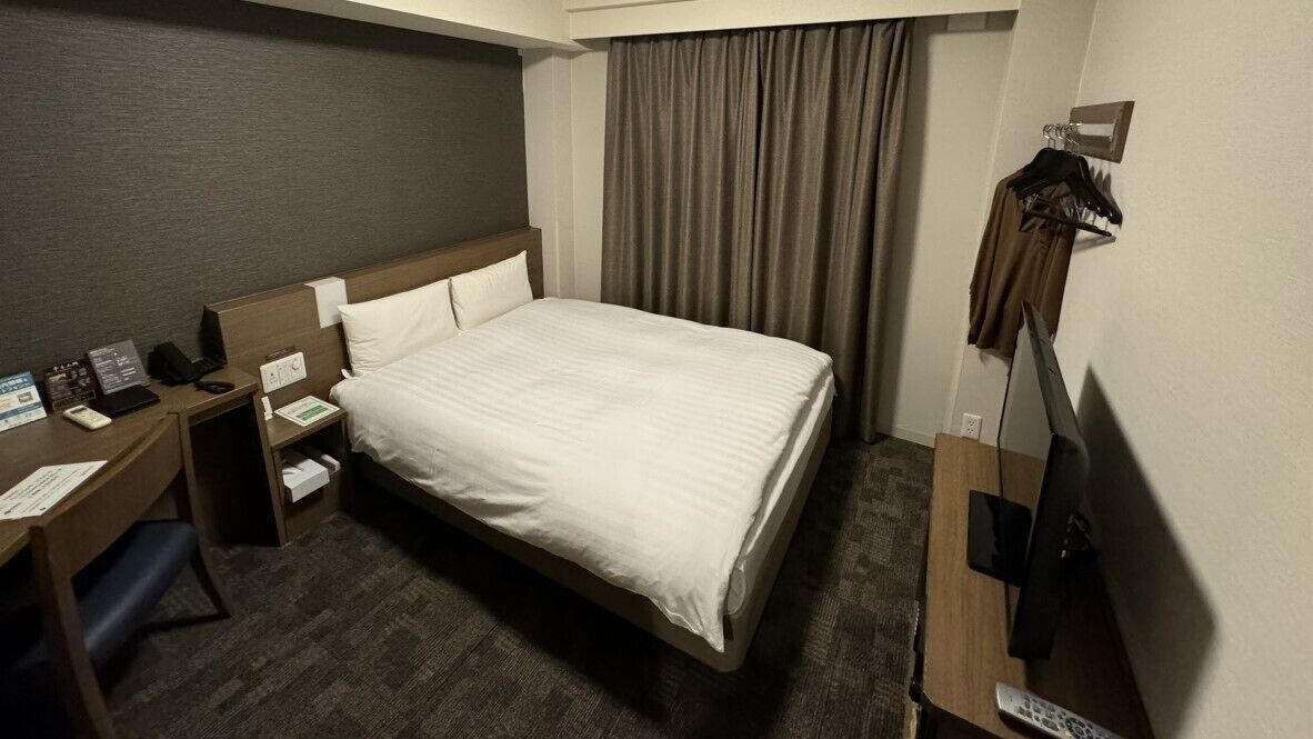 Una habitación de hotel con cama, un escritorio con computadora, un televisor y un perchero.