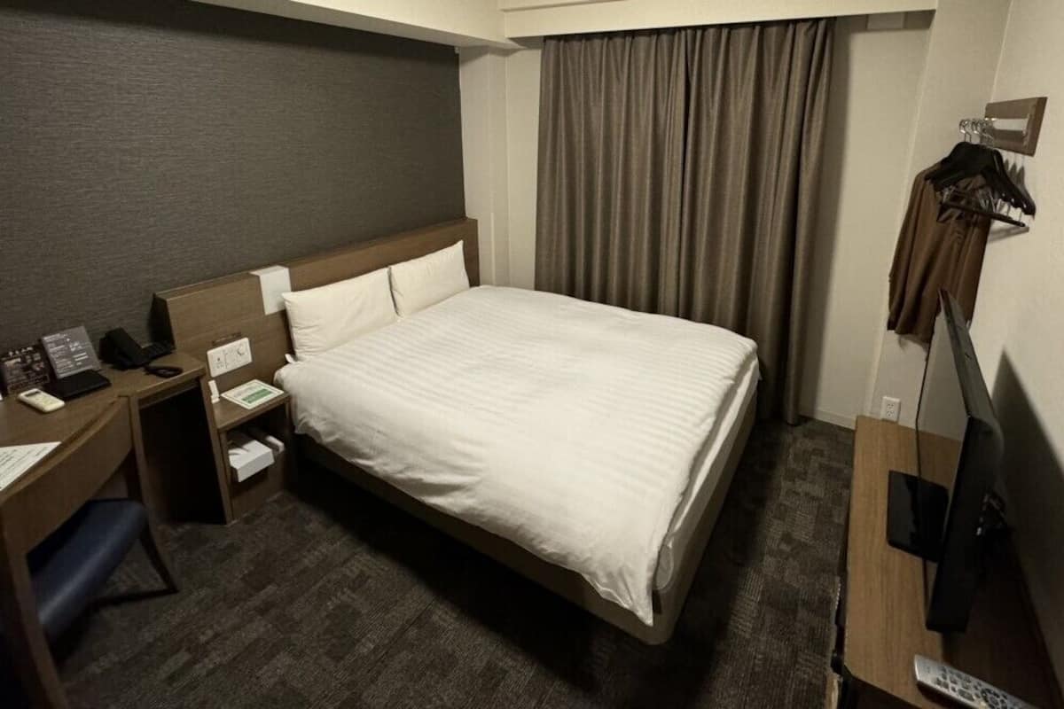 A hotel room with a bed, a desk with a computer, a TV, and a coat rack.