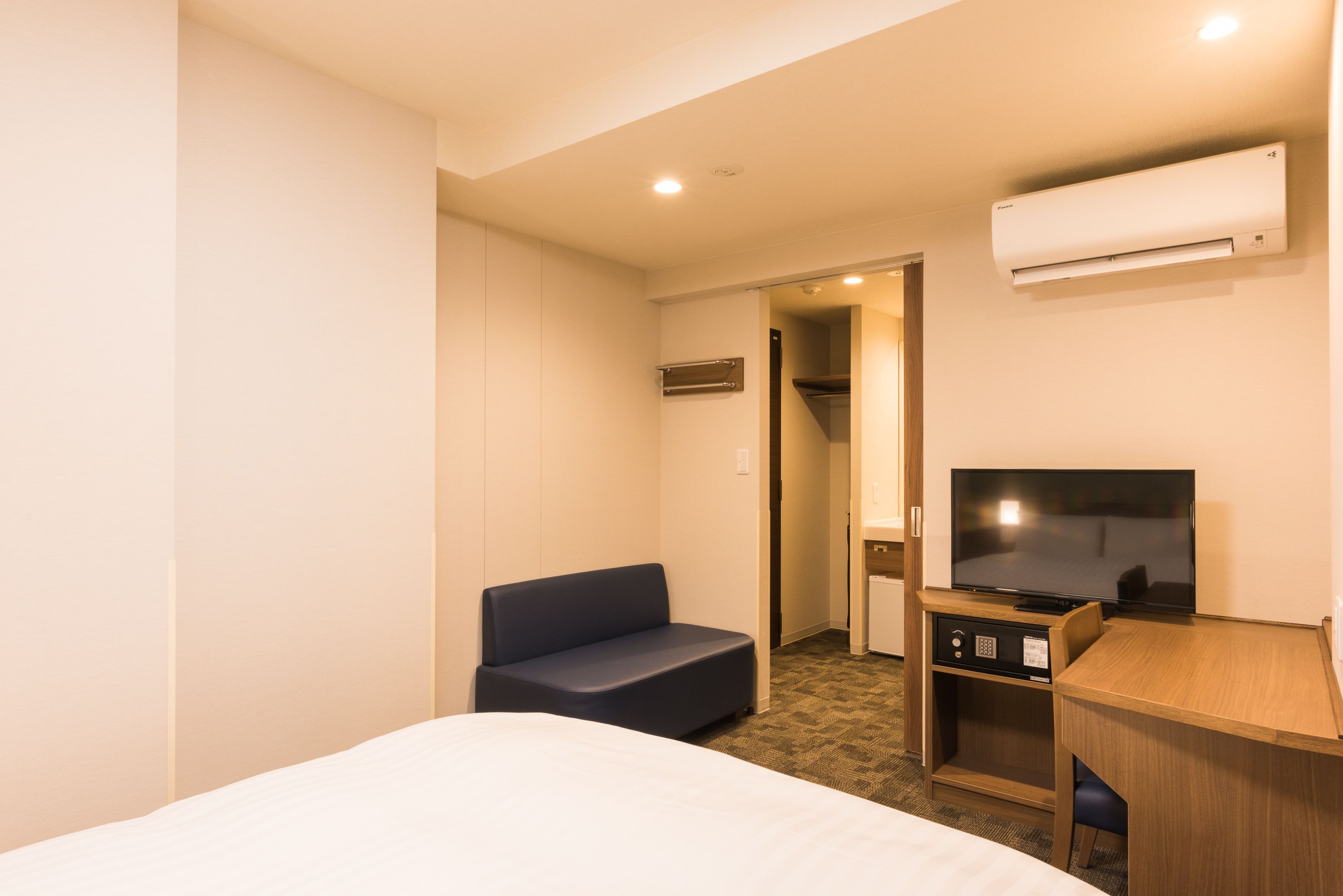A hotel room with a bed, a blue sofa, a desk with a television, and an air conditioning unit.