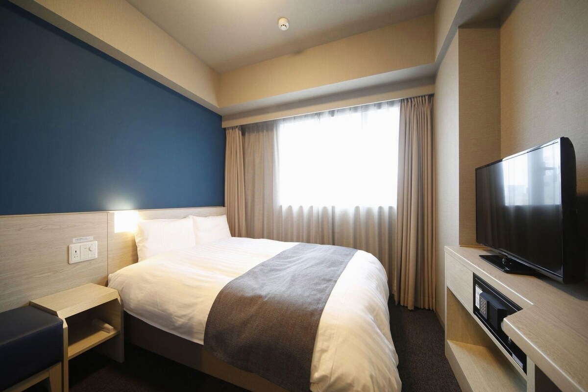 Una habitación de hotel con cama, mesita de noche, televisión y ventana con cortinas.