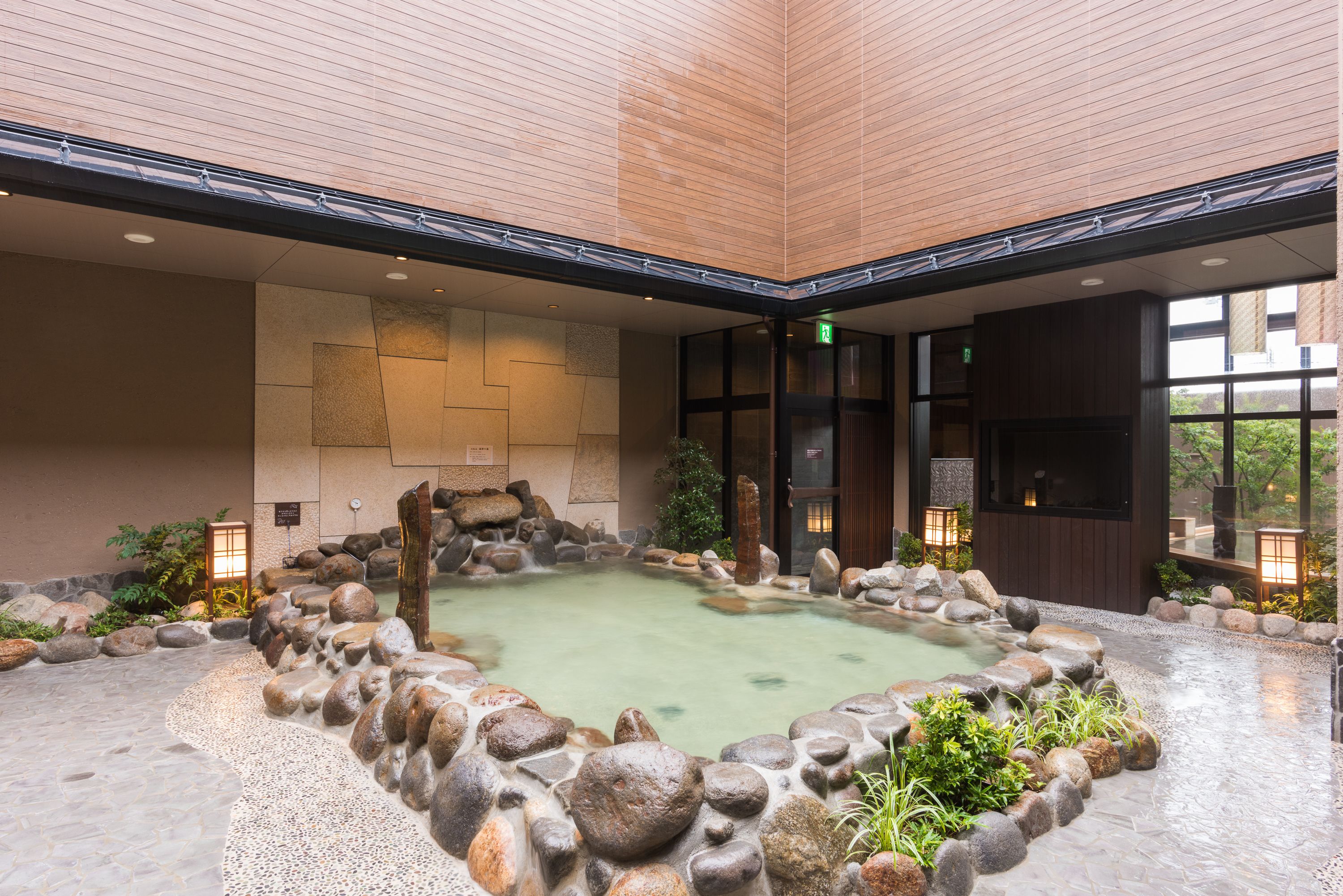 Ein traditionelles japanisches Onsen mit einem Steingrundbecken, umgeben von Steinen und Pflanzen, Holzwänden und einem gefliesten Boden.