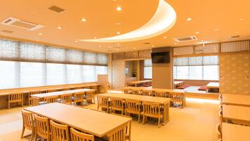 Un vaste espace dédié à la salle à manger, avec de nombreuses tables et chaises en bois, de grandes fenêtres équipées de stores et un plafond au design incurvé.