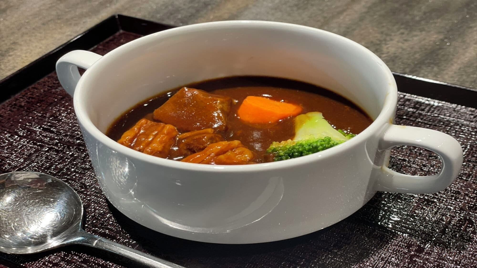A white ceramic mug containing a dark brown soup with chunks of meat, carrots, and broccoli, placed on a textured black tray with a spoon beside it.