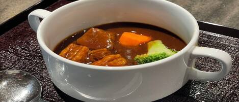 A white ceramic mug containing a dark brown soup with chunks of meat, carrots, and broccoli, placed on a textured black tray with a spoon beside it.