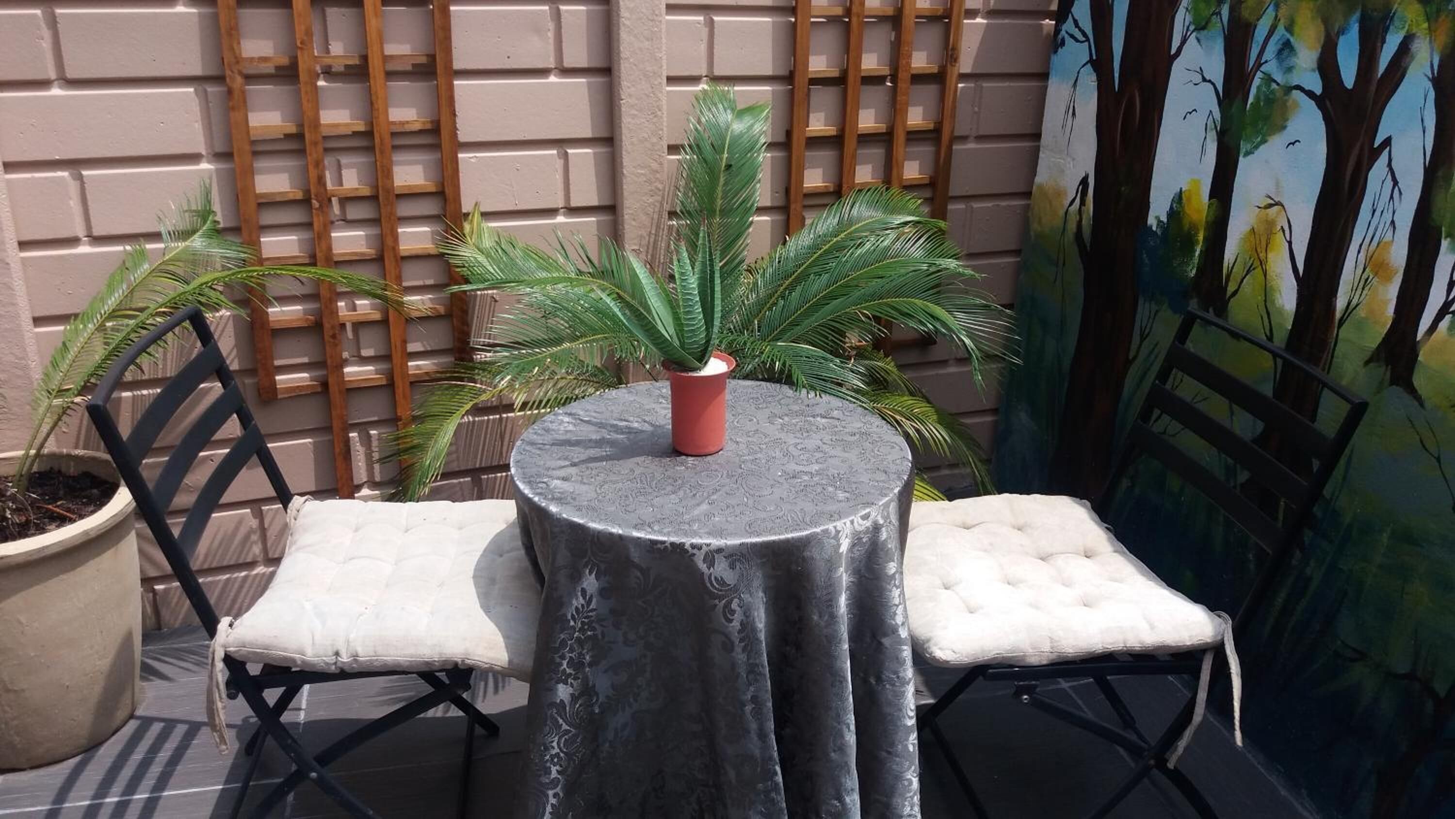 Un petit espace extérieur aménagé avec deux chaises et une table, un pot de fleurs et une fresque murale.