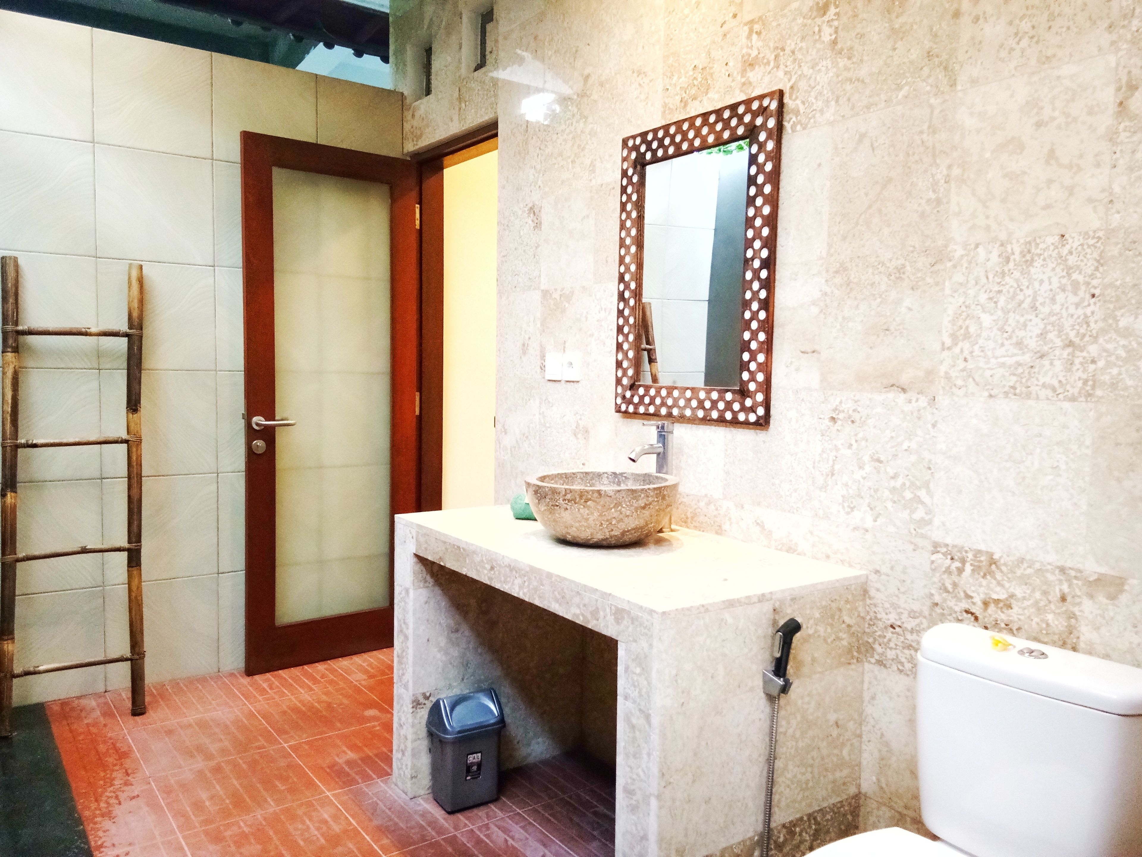 Une salle de bain avec un sol carrelé, une porte en bois, un lavabo avec un receveur en pierre, des toilettes et un miroir avec un cadre à pois.
