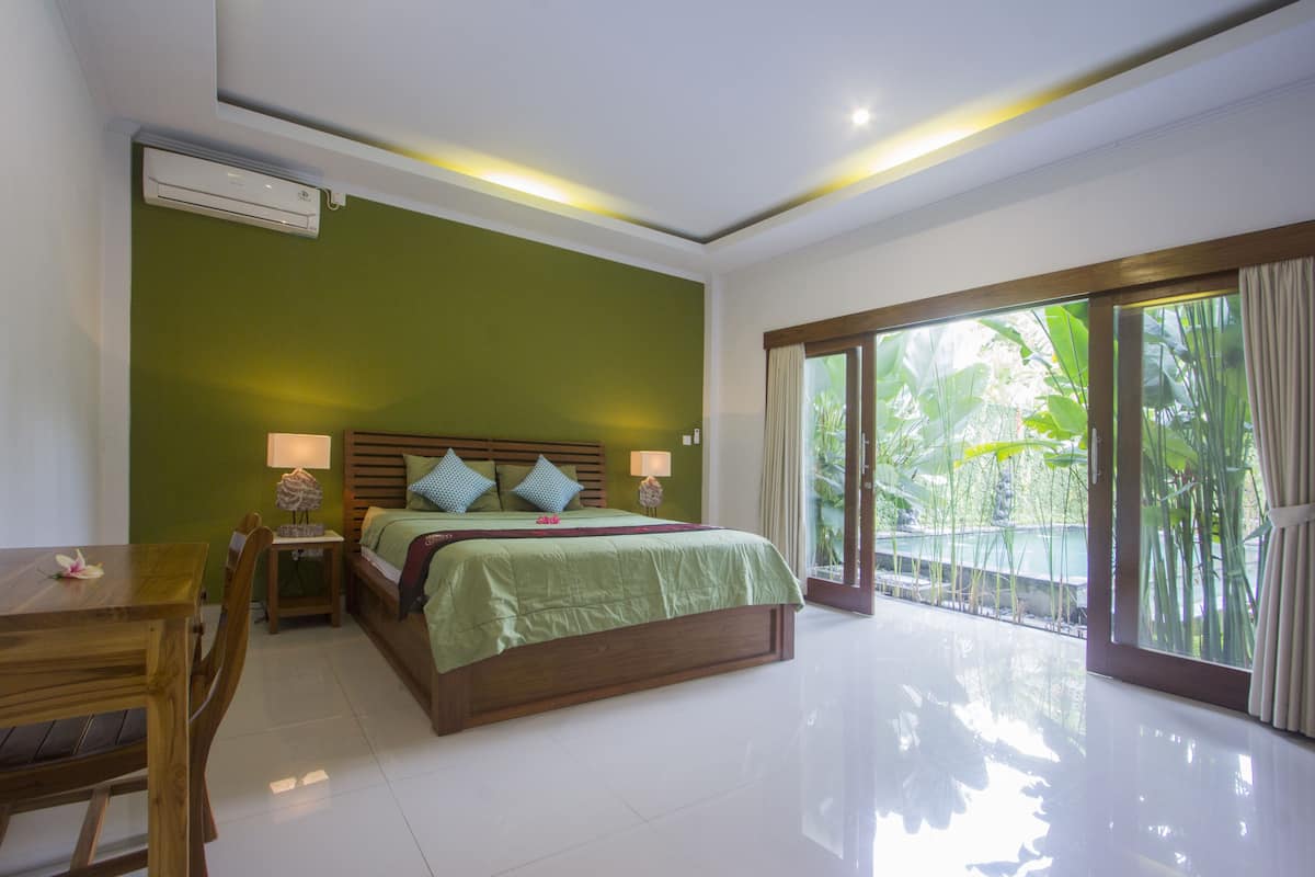 A bedroom with a bed, bedside tables, a wooden desk, and a large sliding glass door leading to a balcony with plants.
