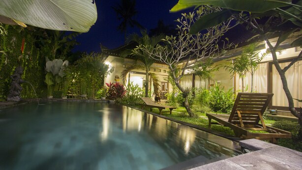 A nighttime view of a swimming pool with a wooden deck and lounge chairs, surrounded by lush greenery and a building with traditional architecture. - Umah Diuma (Ubud)
