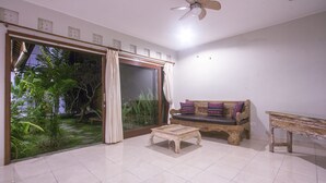 A room with a sliding glass door leading to a garden, a wooden bench with cushions, a small table, and a ceiling fan. - Umah Diuma (Ubud)