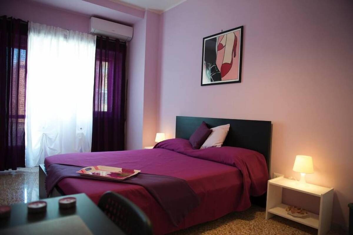 Una habitación de hotel con una cama, dos mesitas de noche con lámparas, un ventanal con cortinas y una obra de arte colgada en la pared.