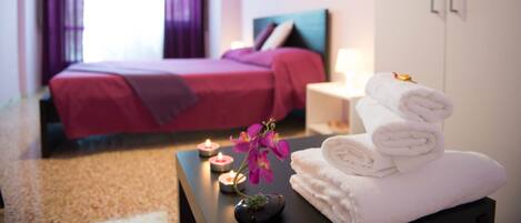 A hotel room with a bed, a nightstand, a table with towels and candles, and a window with curtains.