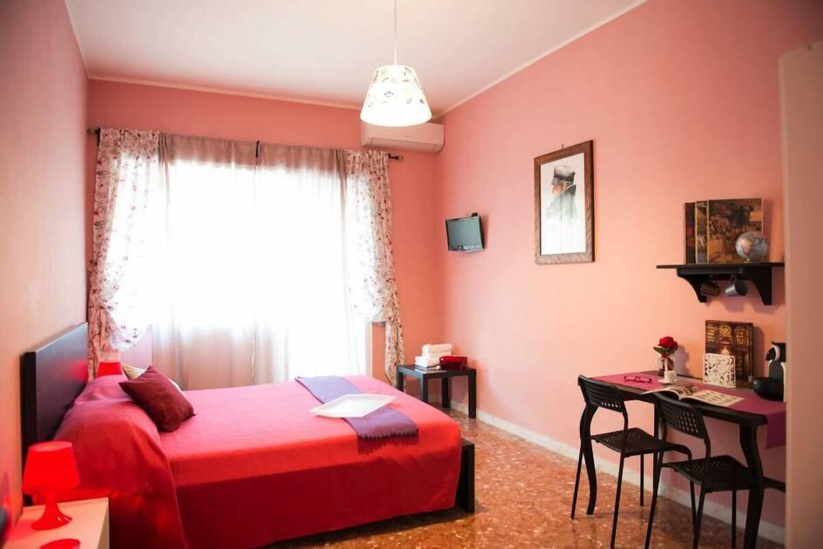 Una habitación con cama, escritorio, sillas, lámpara, televisión y ventana con cortinas.