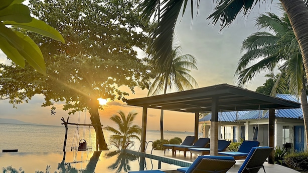 Una piscina infinita con sillones de descanso, palmeras y vistas al mar al atardecer.