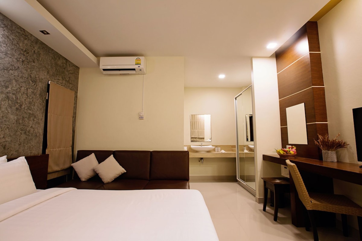 A hotel room with a bed, a sofa, a desk with a chair, a bathroom with a mirror, and an air conditioning unit.