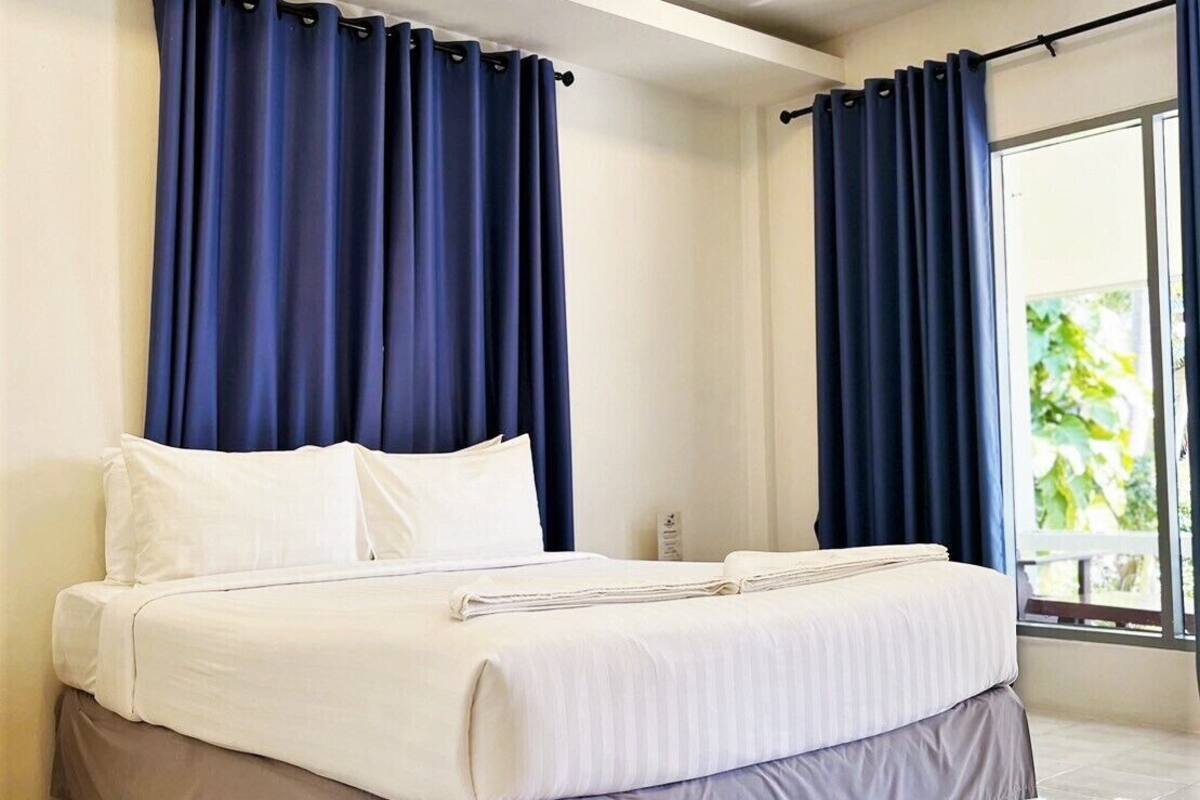 A modern hotel room with a large bed, dark curtains, and a view of greenery through the window.