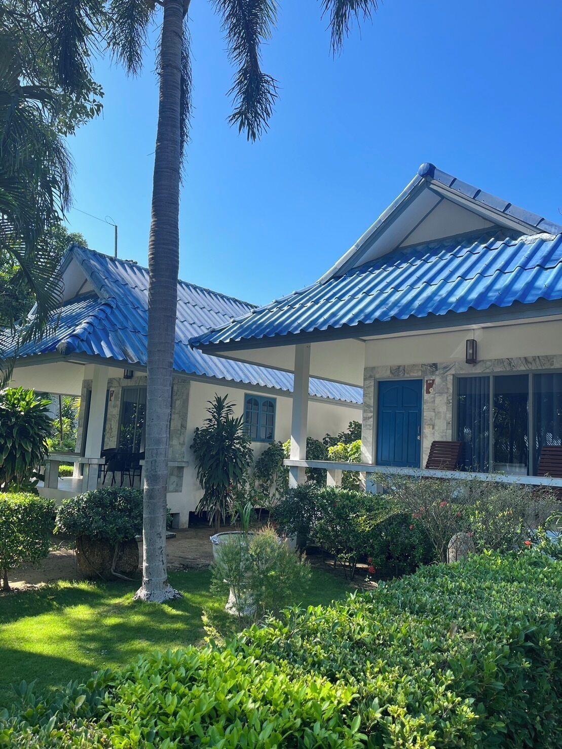 A two-story house with a blue roof, a covered porch, and a well-maintained garden with various plants and a palm tree.