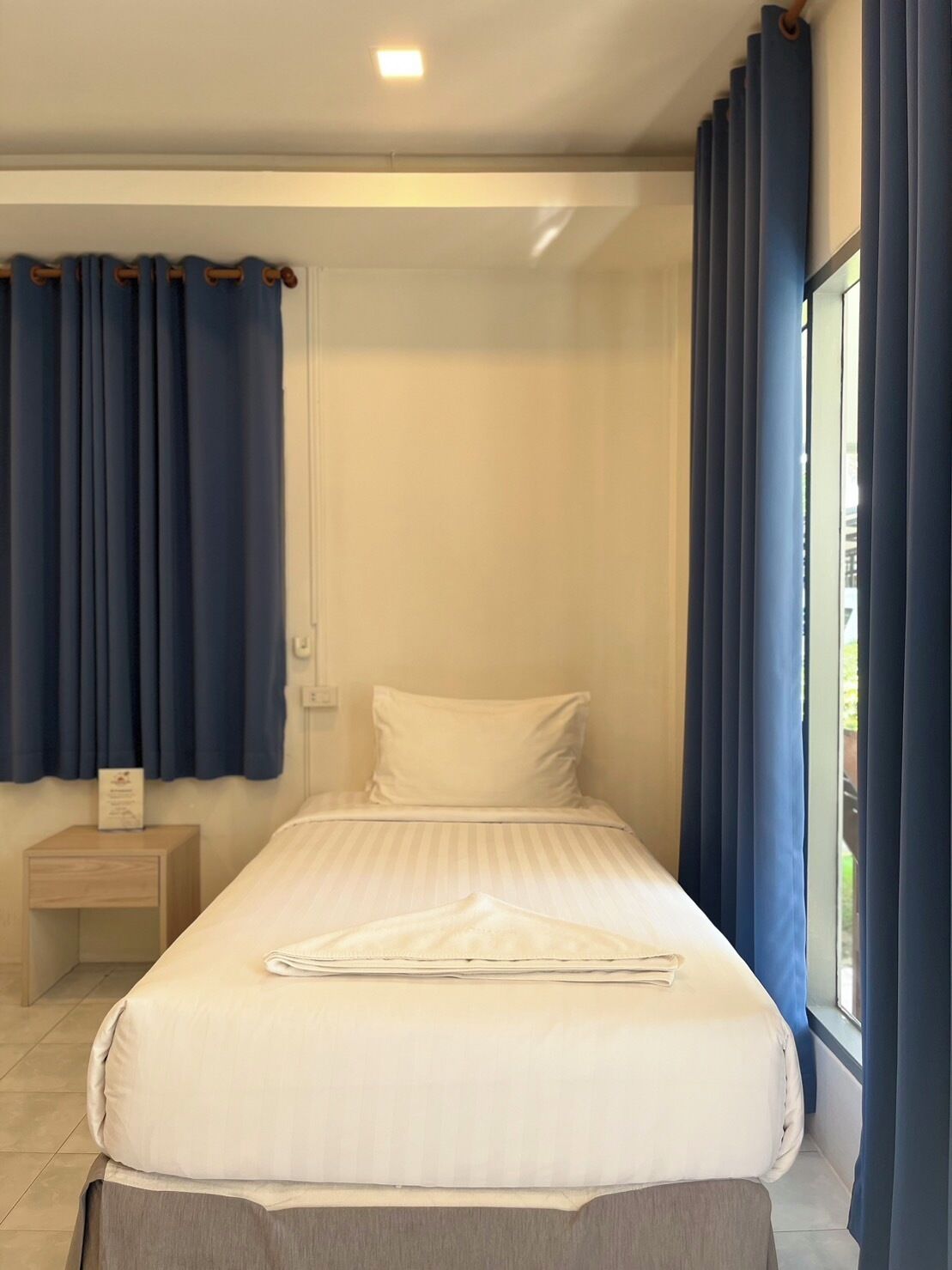 Cama bem arrumada com lençóis brancos, mesa de cabeceira de madeira com um objeto em cima e janela com cortinas azuis.