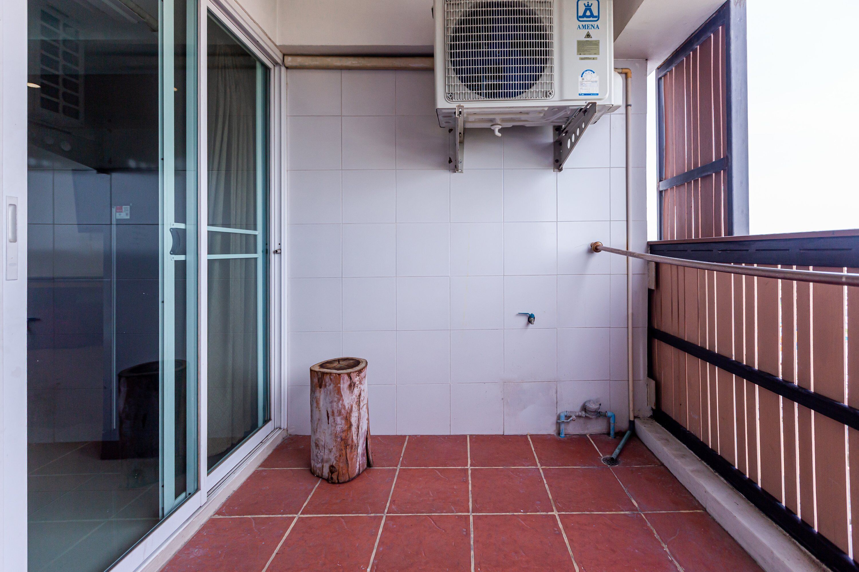 A small balcony with red tiles, a glass door, a metal trash bin, and a white air conditioning unit.