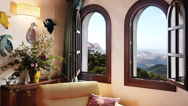 Una habitación con vistas a un paisaje montañoso a través de amplias ventanas de arco, un sofá con un cojín y una lámpara fijada a la pared.