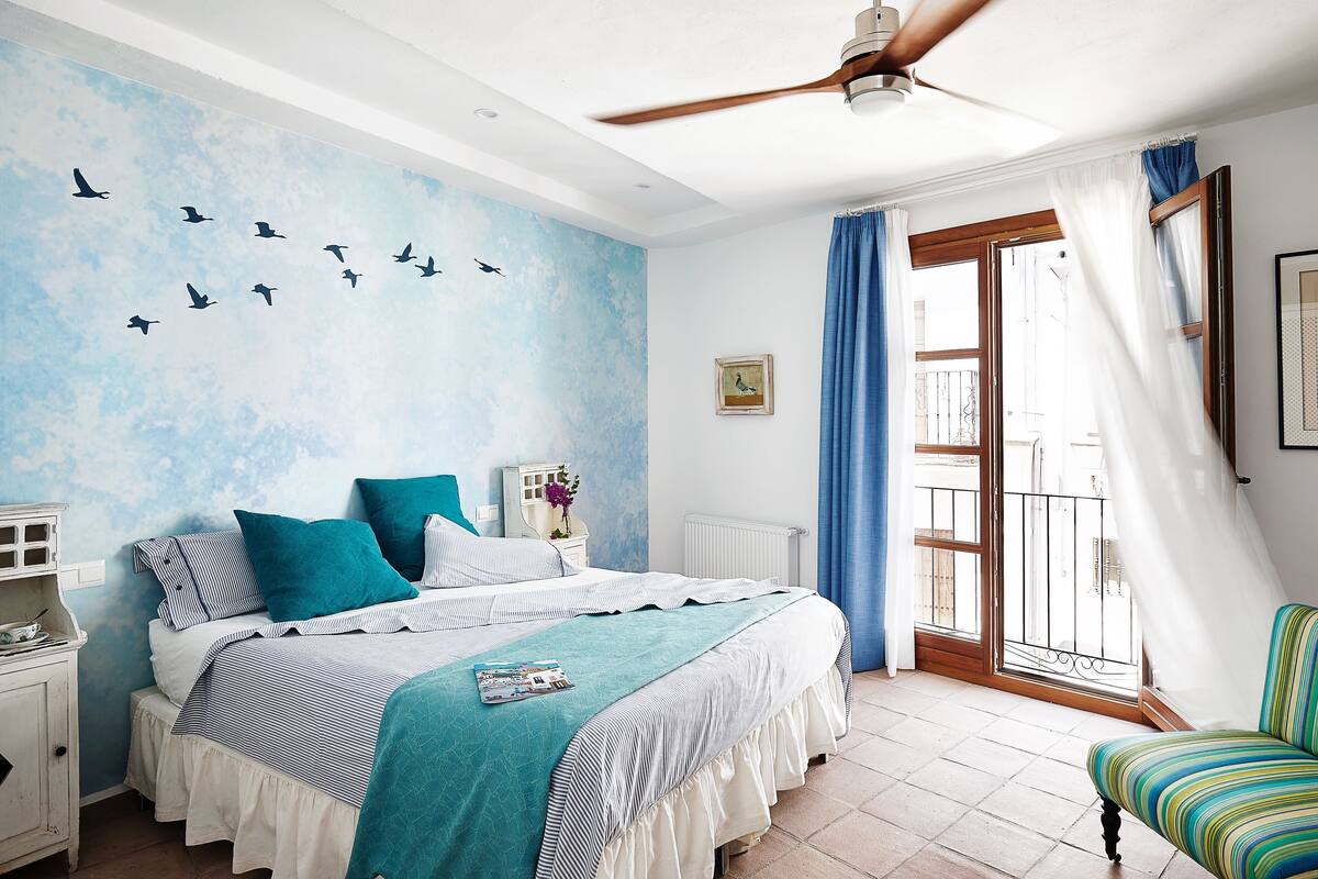 A bedroom with a bed, a nightstand, a chair, a ceiling fan, and a window with curtains.