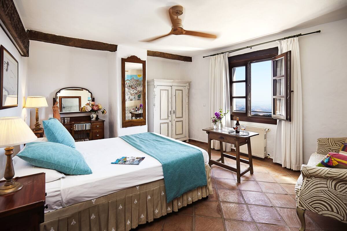 A bedroom with a bed, a desk, a chair, a window with curtains, a ceiling fan, and a view of the outside.