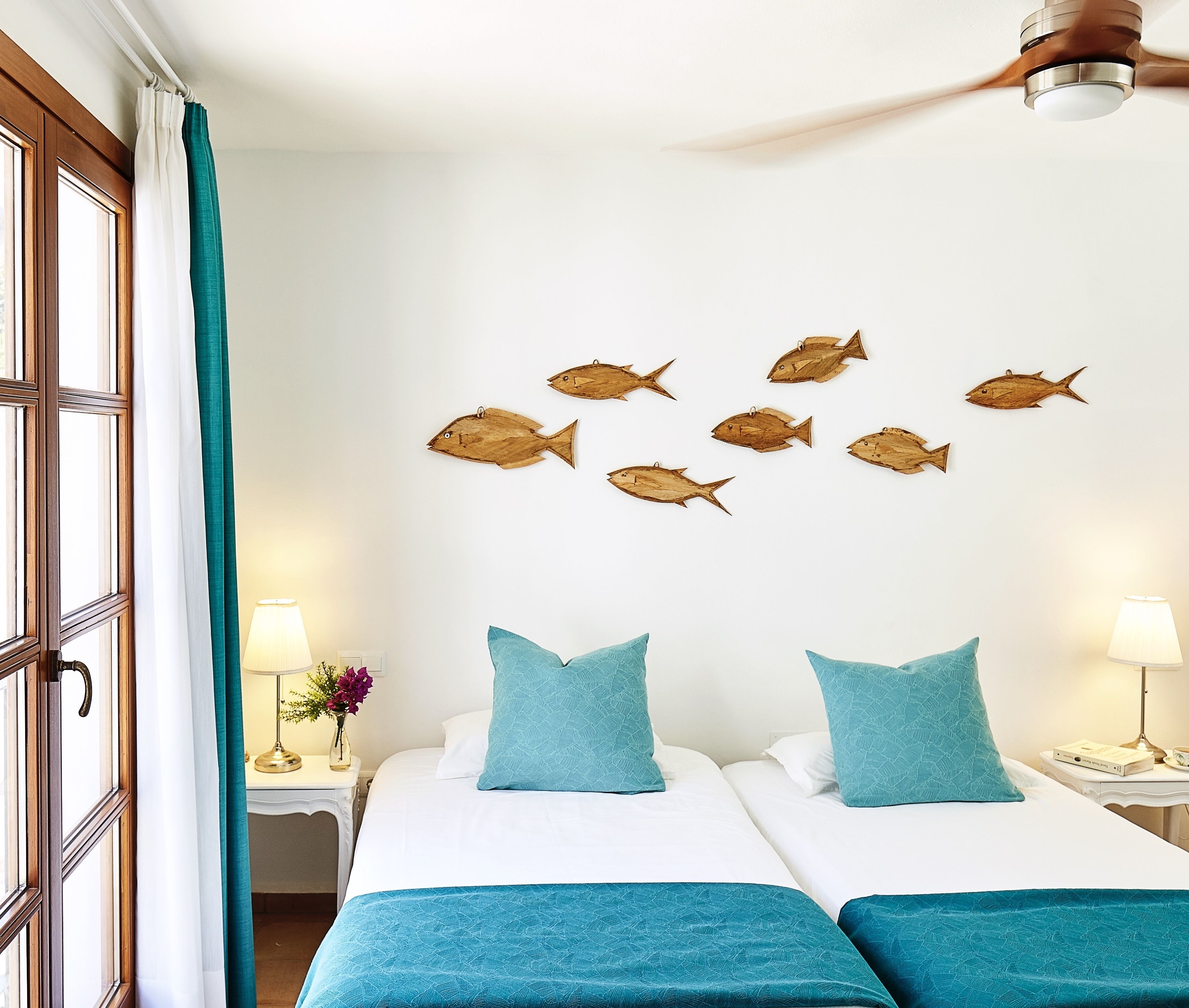 A hotel room with two beds, teal bedding, wooden fish wall art, and a ceiling fan.