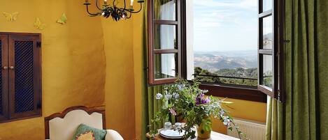 Une pièce donnant sur un paysage grâce à une fenêtre ouverte, une table en bois avec un vase de fleurs, une chaise avec un coussin et un lustre.